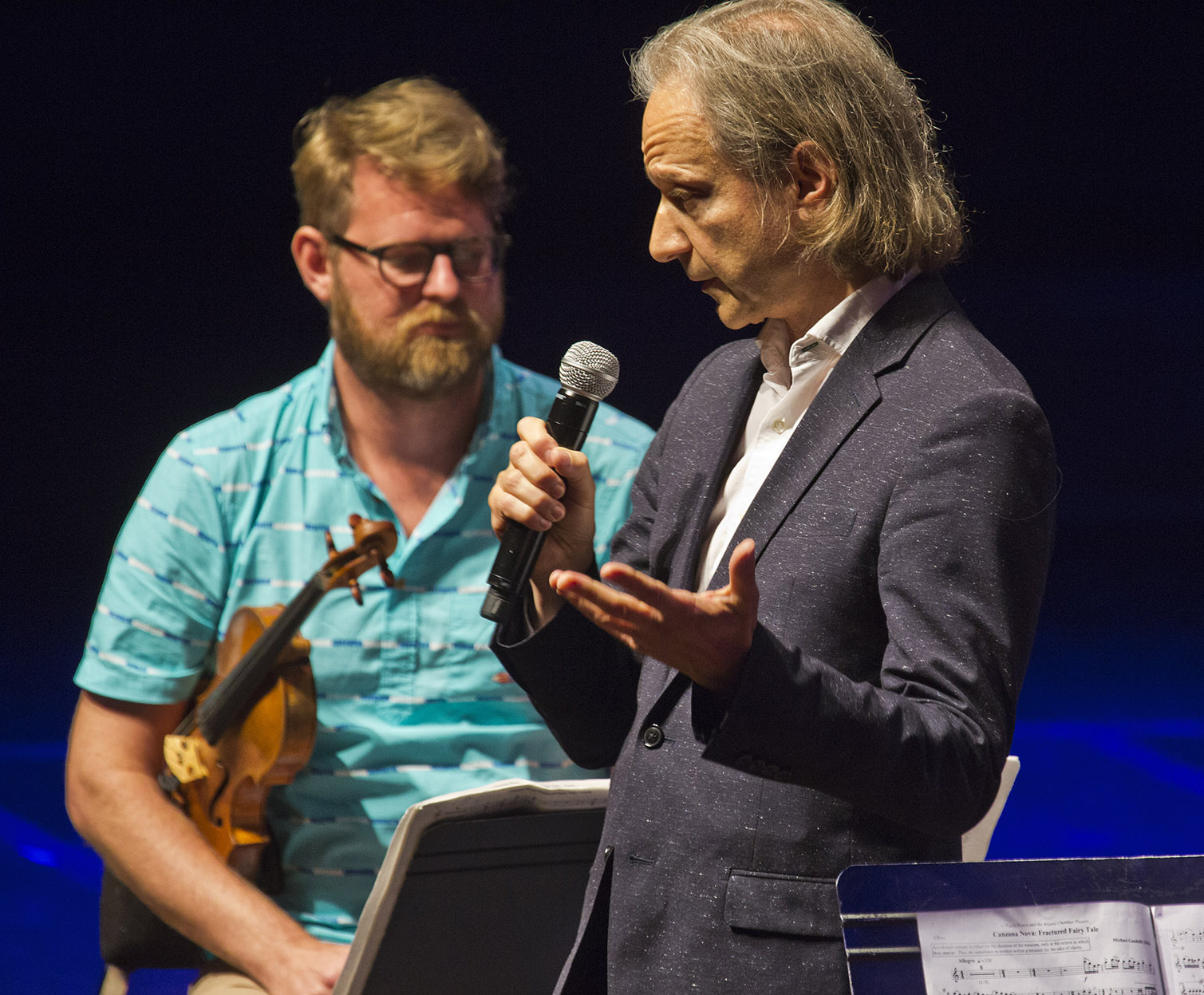 Man speaking into microphone with a violinist in the background